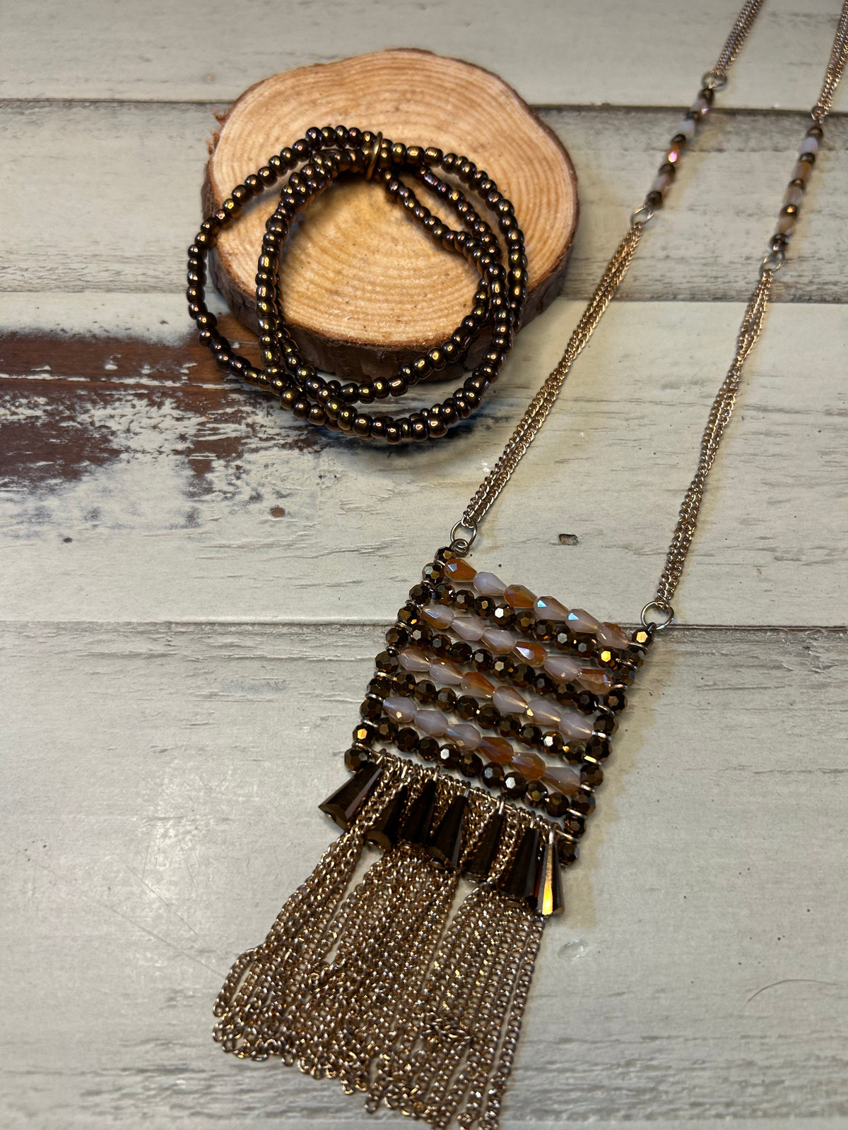 A bohemian-style beaded necklace with a tassel and chain, displayed on a rustic white-washed wood background next to a small wooden slice coaster holding a matching beaded strand.