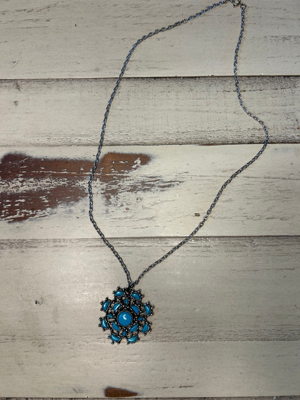 A bohemian-style necklace featuring a dark metal chain and a central snowflake pendant with turquoise blue stones, displayed on a rustic, whitewashed wooden surface.