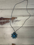 A bohemian-style necklace featuring a dark metal chain and a central snowflake pendant with turquoise blue stones, displayed on a rustic, whitewashed wooden surface.