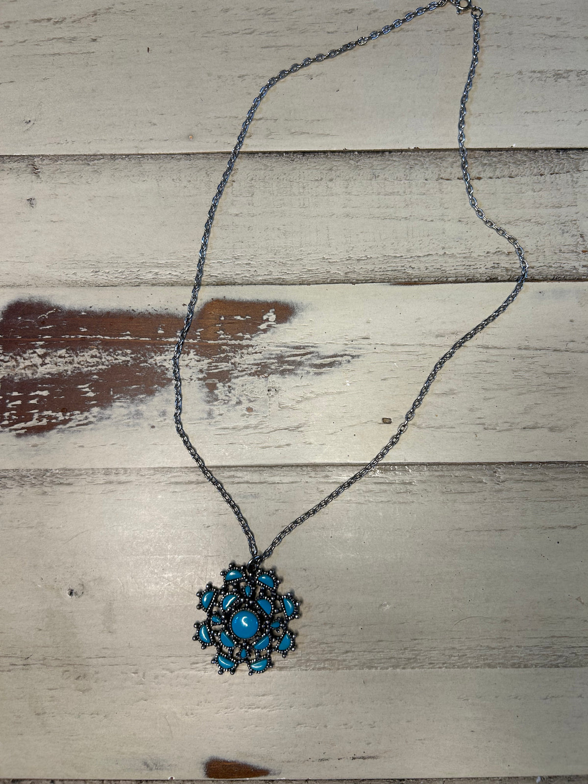 A bohemian-style necklace featuring a dark metal chain and a central snowflake pendant with turquoise blue stones, displayed on a rustic, whitewashed wooden surface.