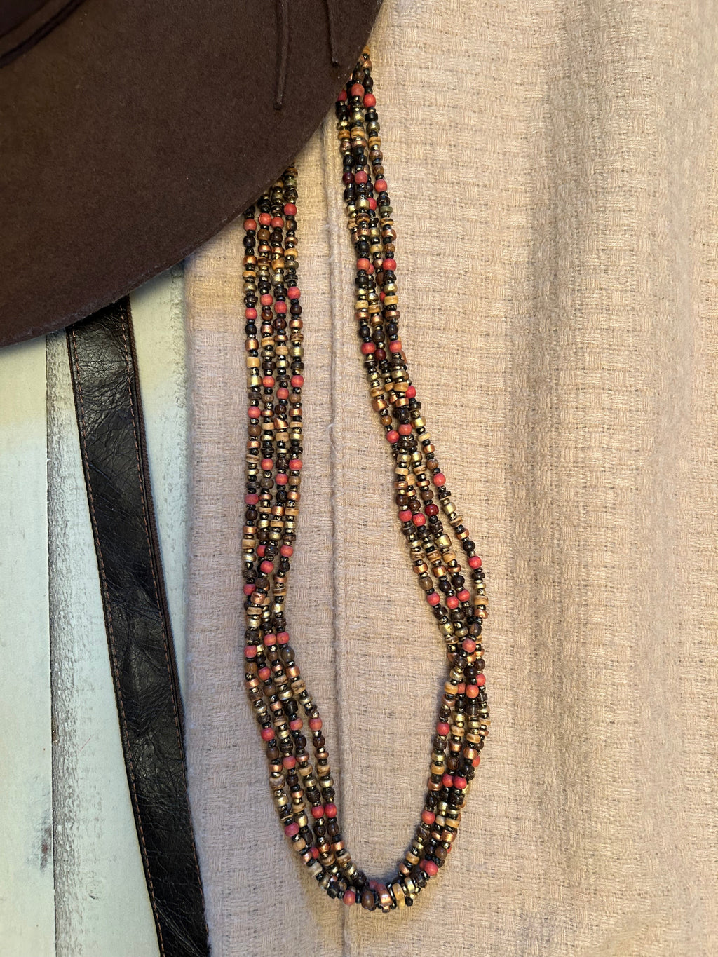 A long beaded belt or strap laid out on a textured beige fabric surface, with a dark brown leather strap and part of a brown hat visible in the background.